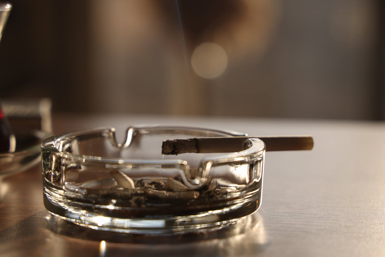A lit cigarette rests on the edge of a clear glass ashtray, with ashes and a faint wisp of smoke visible—a subtle nod to the things hotels used to offer guests. The background is softly blurred, and warm sunlight illuminates the scene.