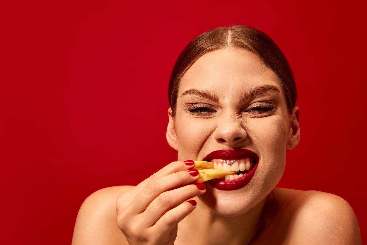 A woman with slicked-back hair and bold red lipstick smiles playfully while holding a french fry near her mouth against a bright red background.