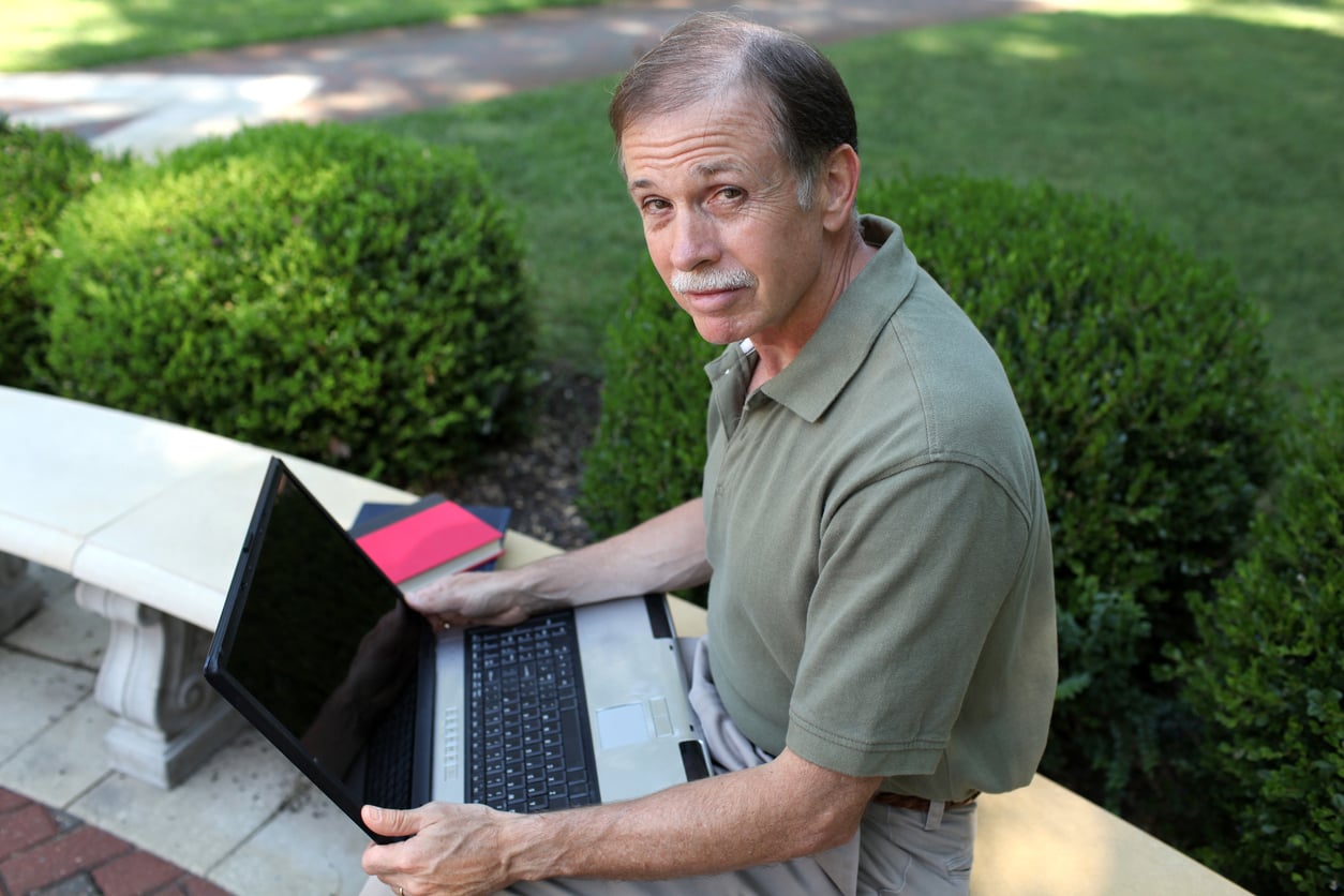 An older man sits on an outdoor bench with a laptop on his lap, looking up and smiling slightly. There are bushes and greenery in the background, and a red notebook beside him.