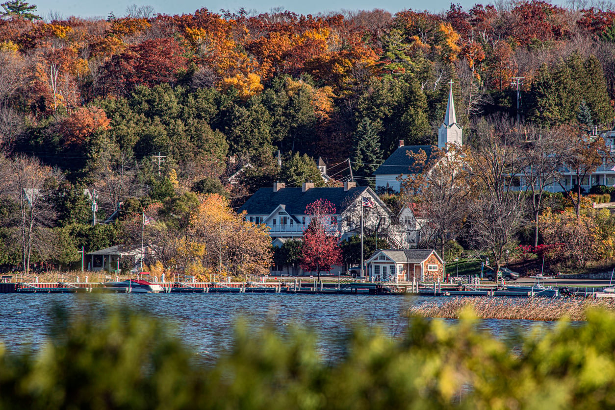 A lakeside village with houses, a white church steeple, and colorful autumn trees—often considered one of the best fall towns in America—is beautifully reflected by the calm water in the foreground.