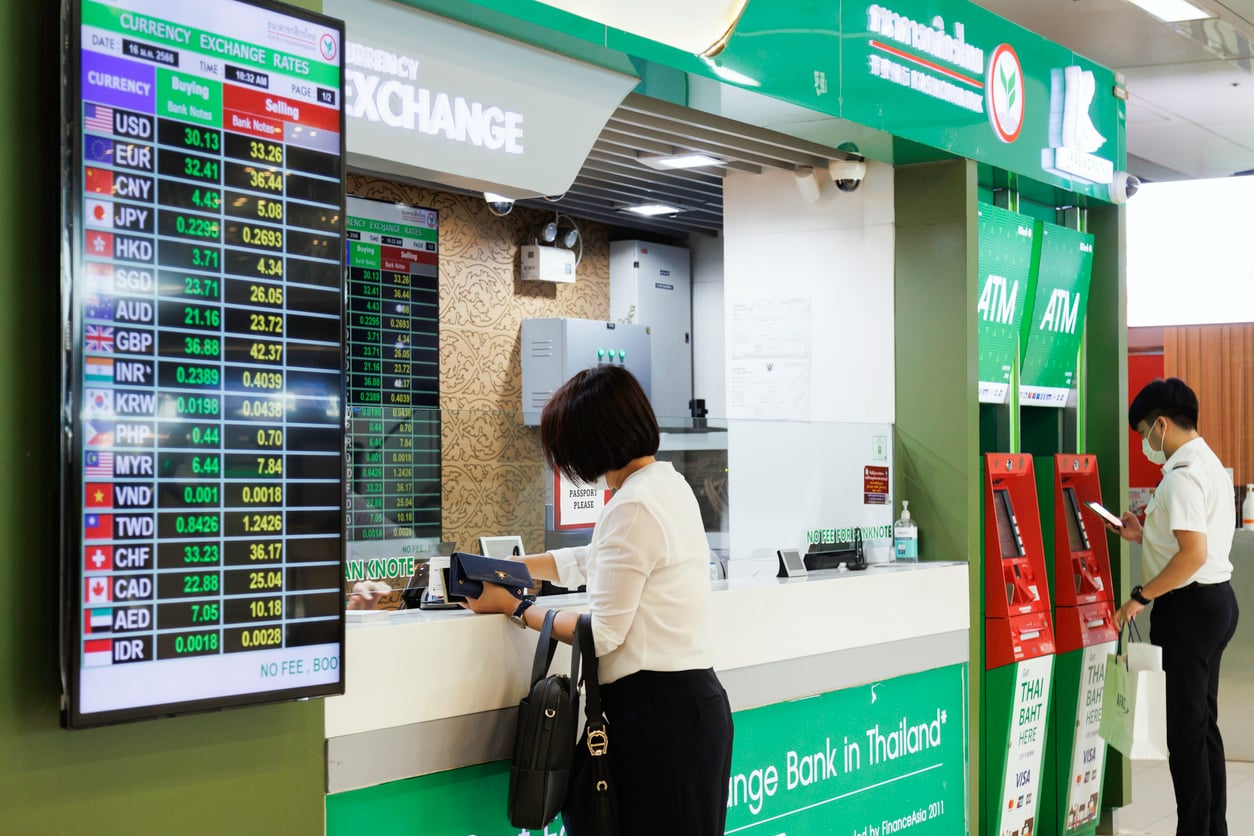 A woman stands at a currency exchange counter next to an electronic board displaying exchange rates, while a man uses nearby ATM machines. Both are inside a brightly lit bank area.
