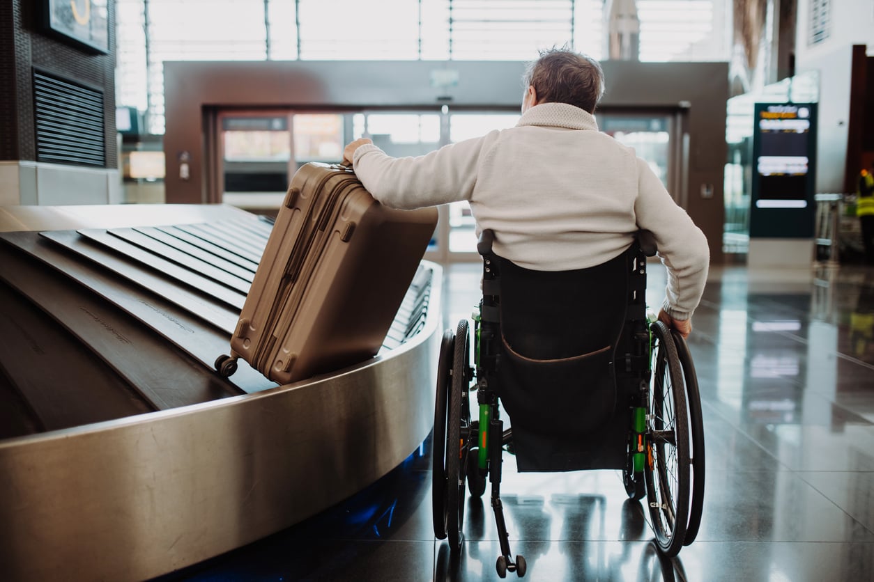 A person in a wheelchair retrieves a suitcase from an airport baggage carousel, seen from behind in a modern, sunlit terminal, highlighting the evolving travel habits of today’s diverse travelers.
