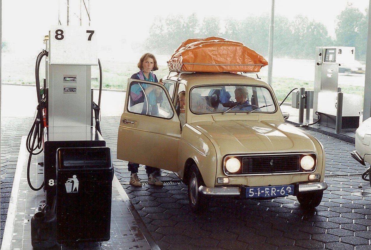 A beige vintage car at a gas station, with a woman standing by the open driver’s door. The car has an orange tarp on the roof and two people inside. Gas pumps are visible in the background.