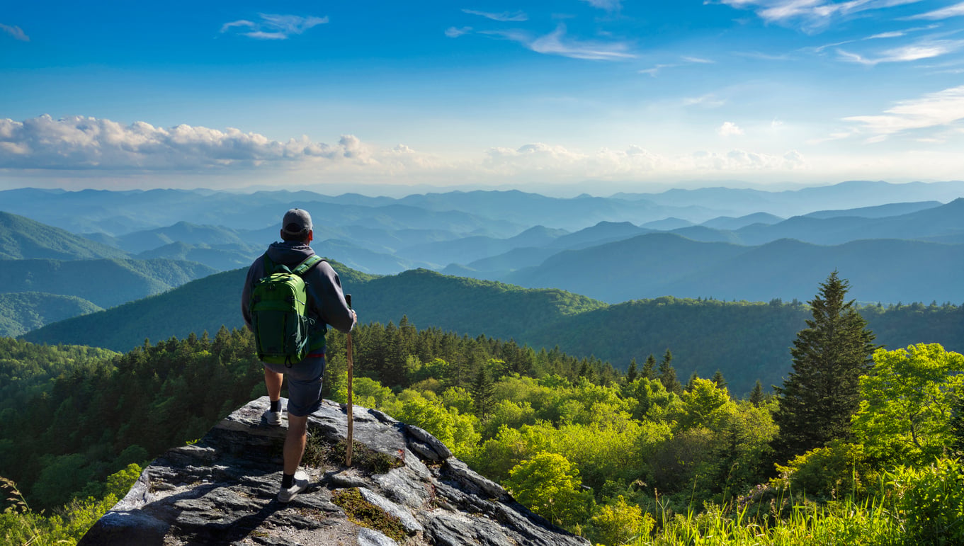 A hiker with a green backpack stands on a rocky outcrop overlooking a vast expanse of lush green mountains under a blue sky with scattered clouds, embracing the spirit of adventure even after age 50.