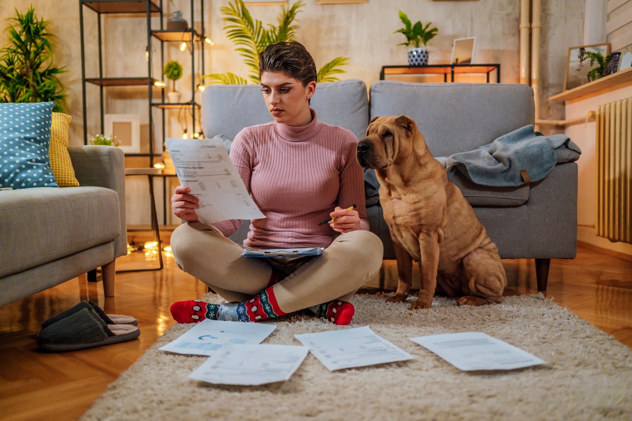 A woman sits on the floor in a warmly decorated living room, reviewing papers with a pen while her dog—clearly well cared for by someone who loves to splurge on their pets—sits faithfully beside her. More paperwork is spread out on the rug in front of them, surrounded by plants and soft lights that add to the cozy atmosphere.