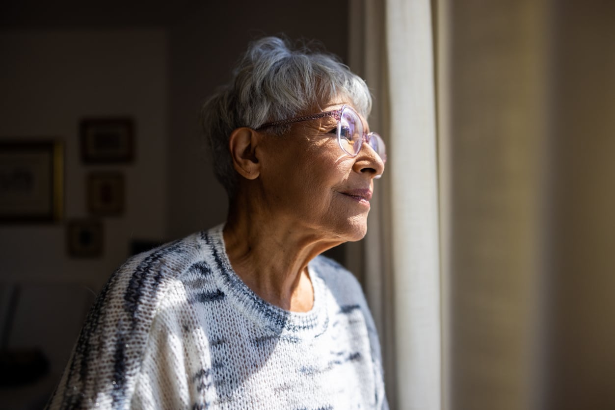 Older woman with short gray hair and glasses gazes thoughtfully out a window, perhaps reflecting on retirement tips she’s gathered over the years. Sunlight gently illuminates her face as she sits in a light patterned sweater. The softly blurred background suggests a calm indoor setting, perfect for contemplation.