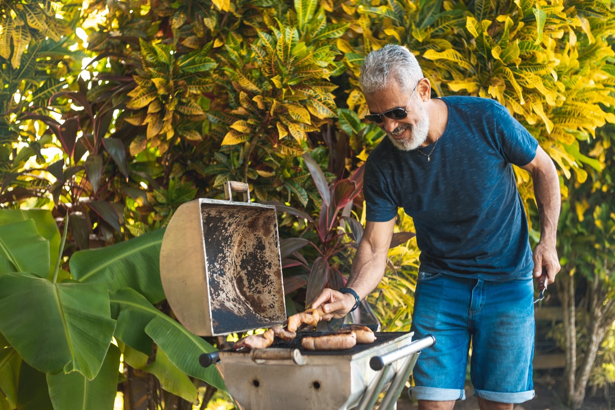 A man with gray hair and a beard, wearing sunglasses, a blue shirt, and shorts, grills sausages outdoors. He is smiling and surrounded by lush green and yellow tropical plants.