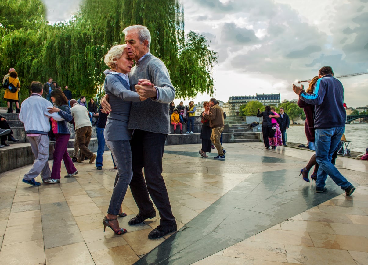 Older couples dance closely in an outdoor plaza by a river, surrounded by trees and other people whose travel behaviors bring them to this vibrant spot. The sky is cloudy, and the setting feels lively and relaxed.