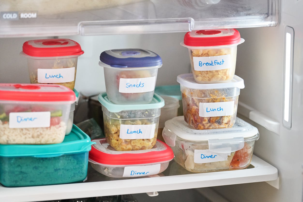 Clear containers with red and blue lids, labeled with “Lunch,” “Dinner,” “Breakfast,” and “Snacks,” are neatly stacked and stored in a refrigerator. Meals inside are visible through the containers.