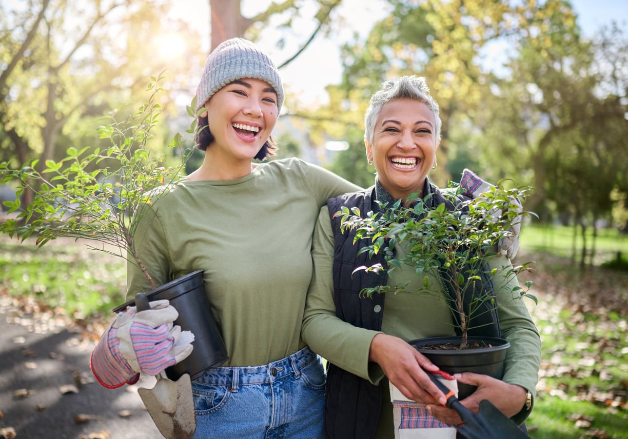 Two women smiling while holding potted plants in a park, sharing retirement tips as they garden together. One wears gloves and a beanie, the other has short hair and pruners. Sunny day, autumn leaves on the ground.