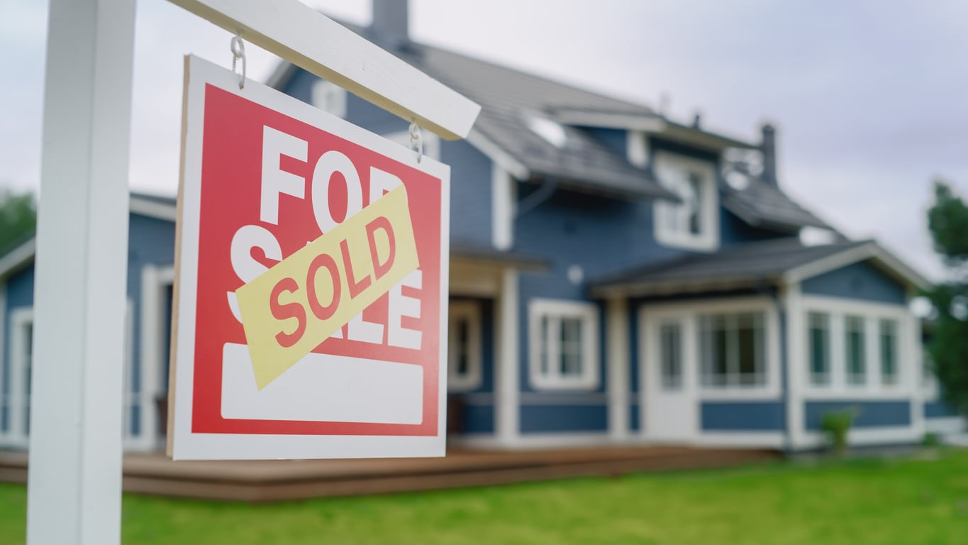 A red “For Sale” sign with a yellow “Sold” sticker is posted in front of a modern blue house with white trim, sitting on a green lawn.