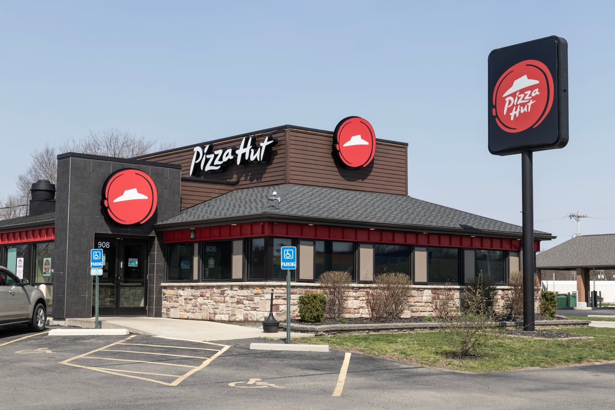 A Pizza Hut restaurant with the red logo on the building and sign, parking lot in front, and two accessible parking spaces visible on a clear day—perfect for discovering some Wild Fast-Food Facts while you dine.