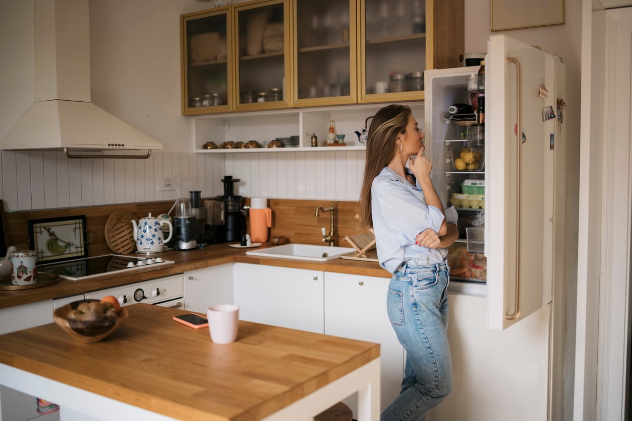 A woman stands in a kitchen, looking thoughtfully into an open refrigerator. The kitchen has white cabinets, wooden countertops, and various appliances and jars displayed on the shelves.
