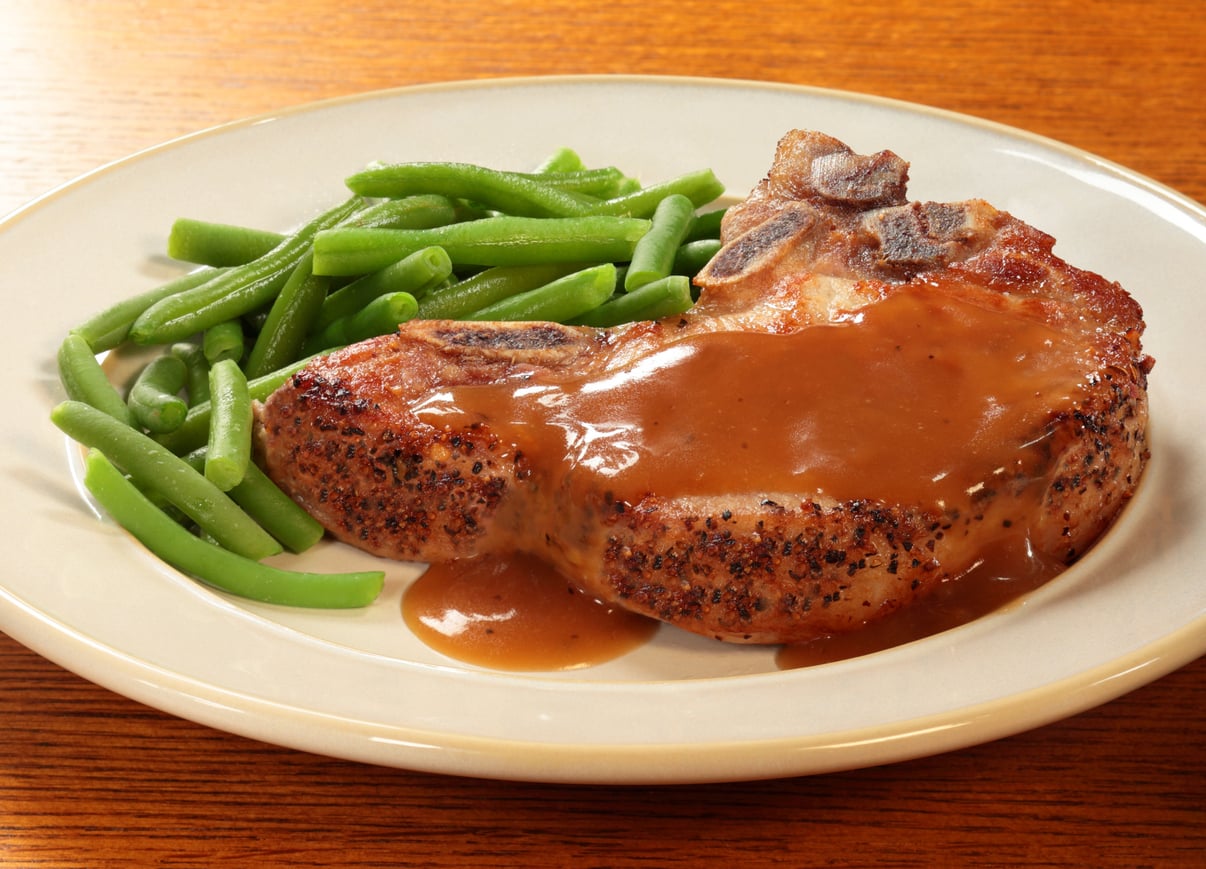 A plate with a seasoned pork chop covered in brown gravy and a side of cooked green beans, served on a light-colored dish with a wooden table background—just like the comforting meals middle-class moms made.