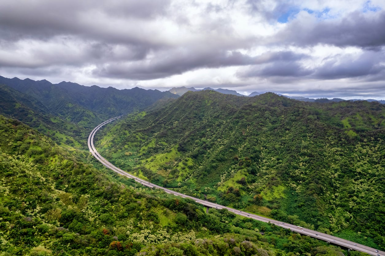 A curving highway winds through lush, green mountains under a cloudy sky, surrounded by dense vegetation and rolling hills.