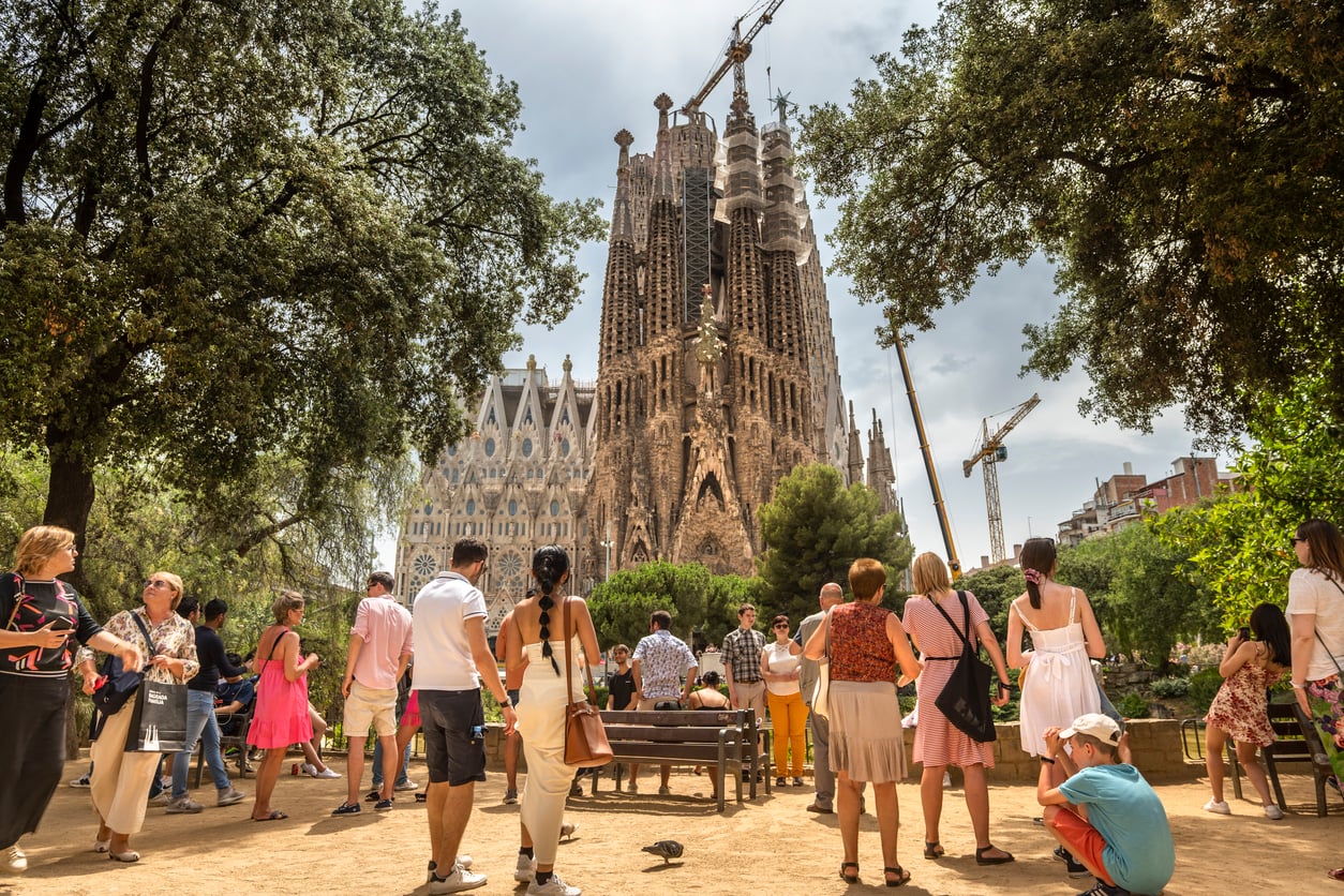 Tourists gather in a sunny park with trees, viewing the Sagrada Família basilica in Barcelona, which is under construction with cranes above its ornate towers.