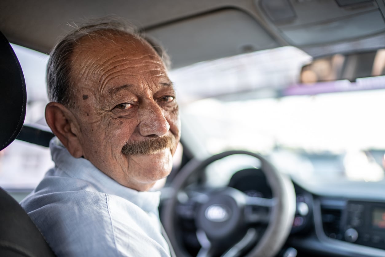 An elderly man with a mustache sits in the driver’s seat of a car, wearing a light blue shirt and smiling at the camera. The car’s steering wheel and dashboard are visible, reflecting how Baby Boomer changes have shaped both his generation’s lifestyles and the vehicles they drive.
