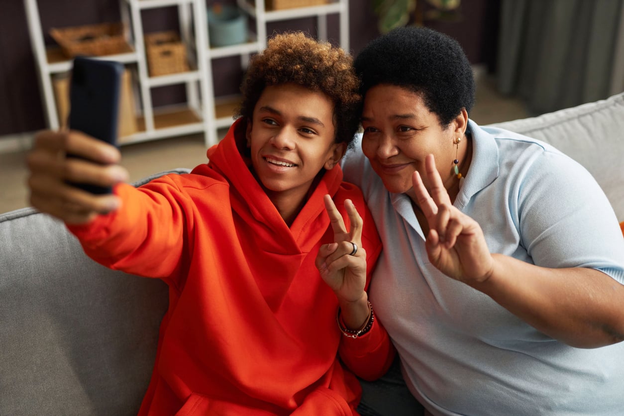 Teen in a red hoodie and adult in a light blue shirt sit on a couch, smiling and making peace signs while taking a selfie together with a smartphone.