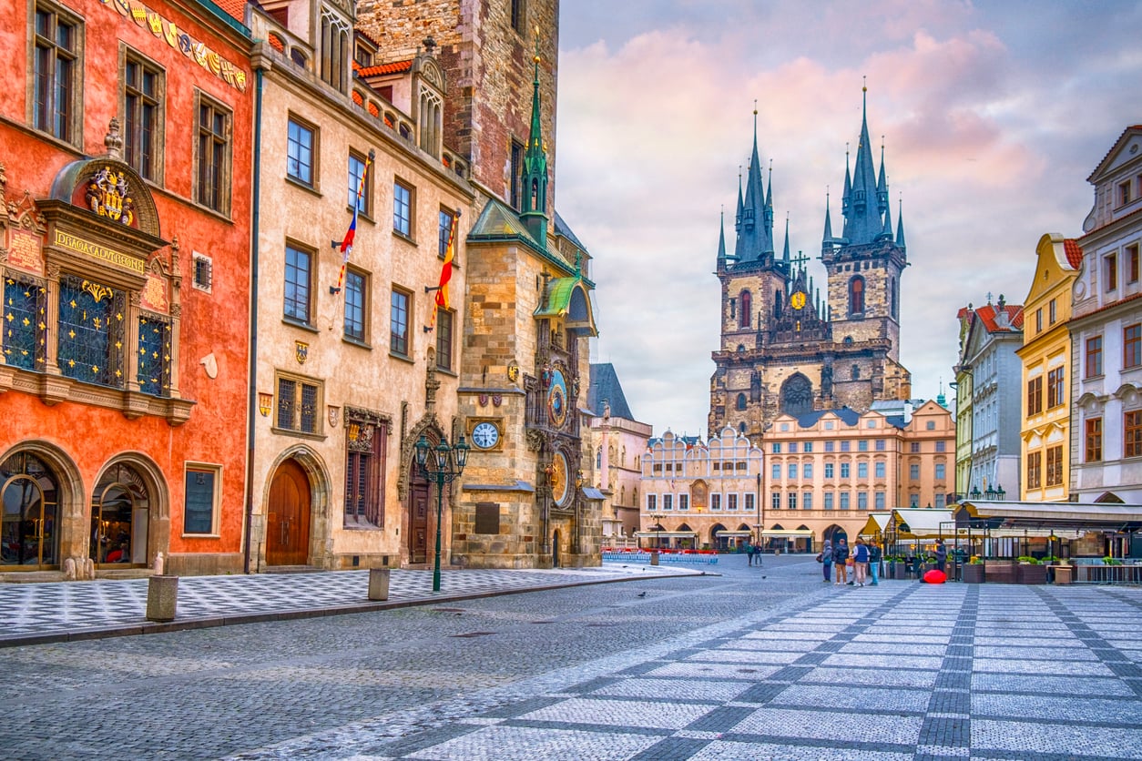 Colorful historic buildings and the Prague Astronomical Clock on Old Town Square, with the Church of Our Lady before Týn in the background, stand beneath a blue and pink sky; a few people stroll across the patterned stone square, likely discussing their experiences with bad food from nearby cafés.