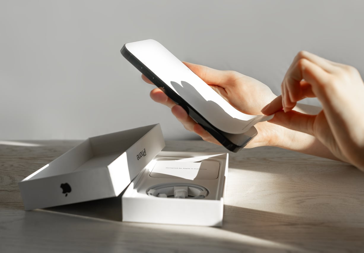 A person peels protective film off a new smartphone next to an open Apple box with charging cable inside, on a sunlit table.