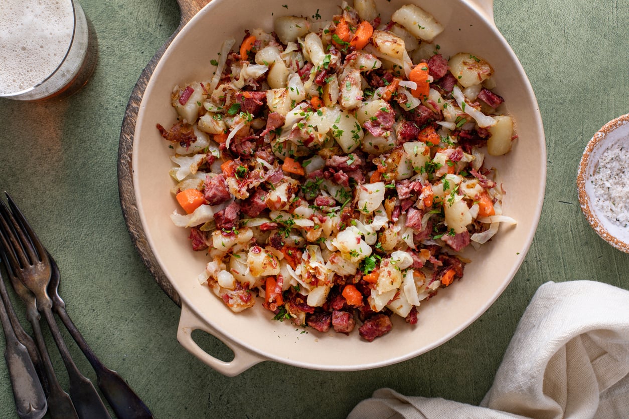 A bowl filled with corned beef hash made of diced potatoes, corned beef, carrots, and herbs sits on a green table next to a drink, utensils, and a white napkin.