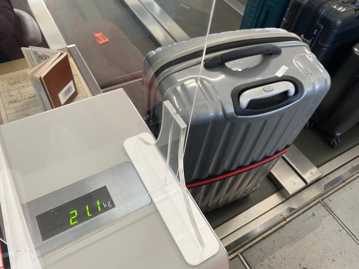 A digital scale displays 21.1 kg next to a silver suitcase on a luggage belt at an airport check-in counter. A passport and boarding pass are partially visible on the left.