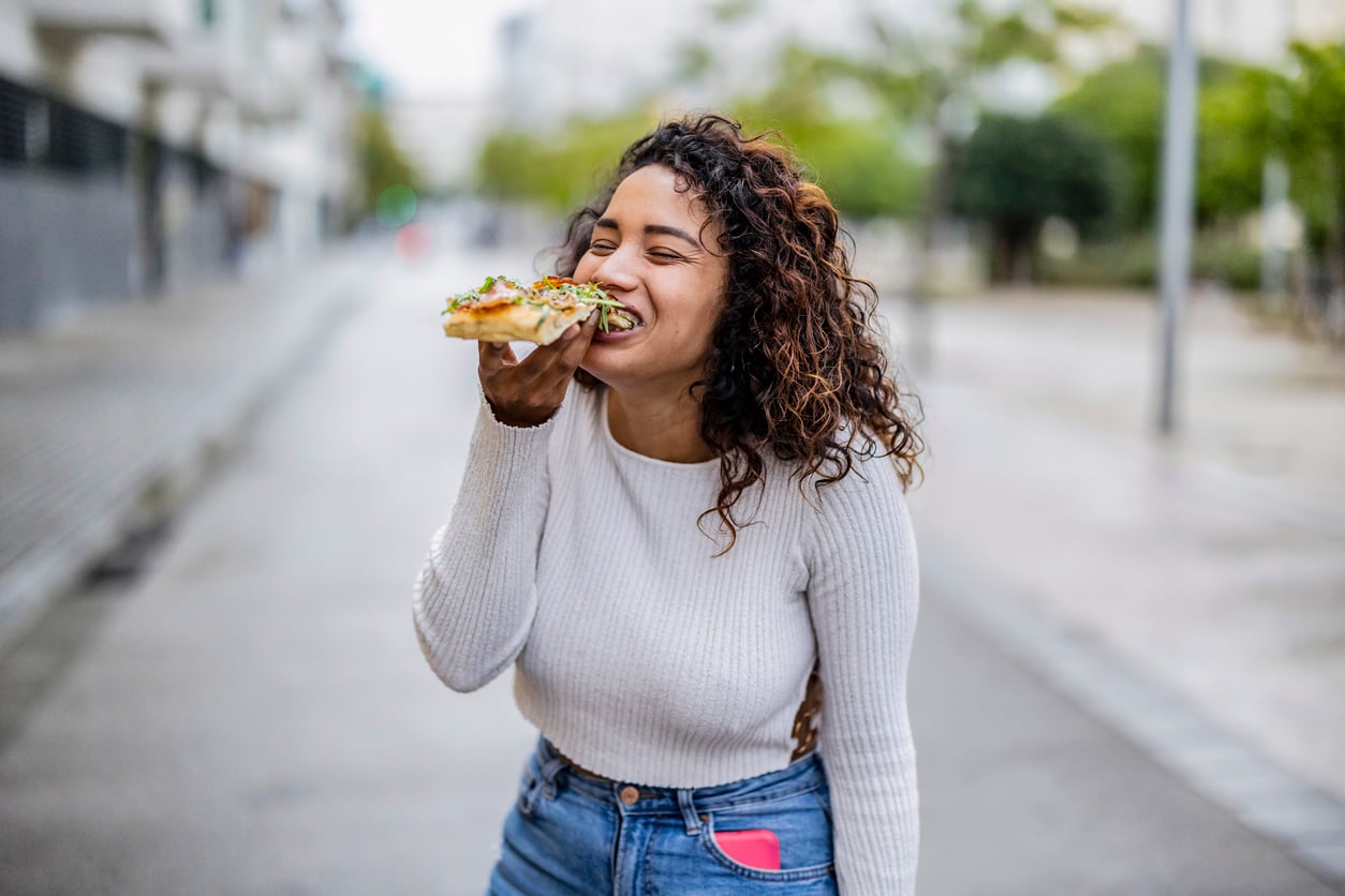 A woman with curly hair laughs while holding and eating a slice of pizza outdoors on a city street. She is wearing a white sweater and blue jeans with a pink phone in her pocket.