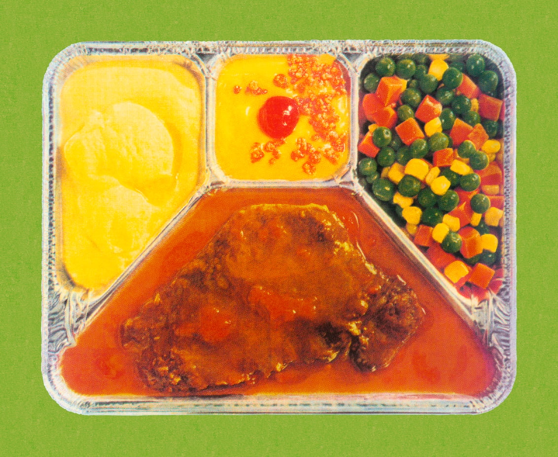 A TV dinner in a divided tray with mashed potatoes, corn with a cherry on top, mixed vegetables, and a piece of meat in brown sauce, all on a green background.