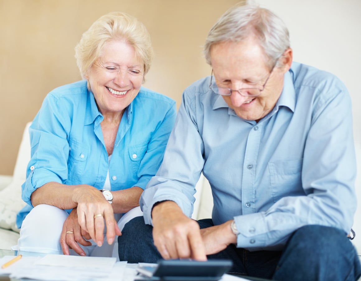 Smiling older couple sitting together, reviewing paperwork and using a calculator at a table, both wearing light blue shirts as they discuss retirement tips.
