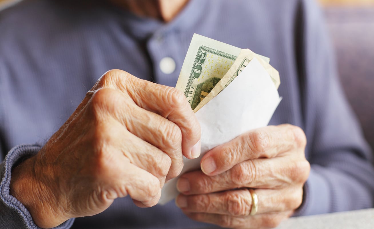 An elderly person wearing a blue shirt holds a white envelope filled with U.S. dollar bills, their hands and the money in clear focus—an image that subtly highlights the importance of retirement tips for financial security.