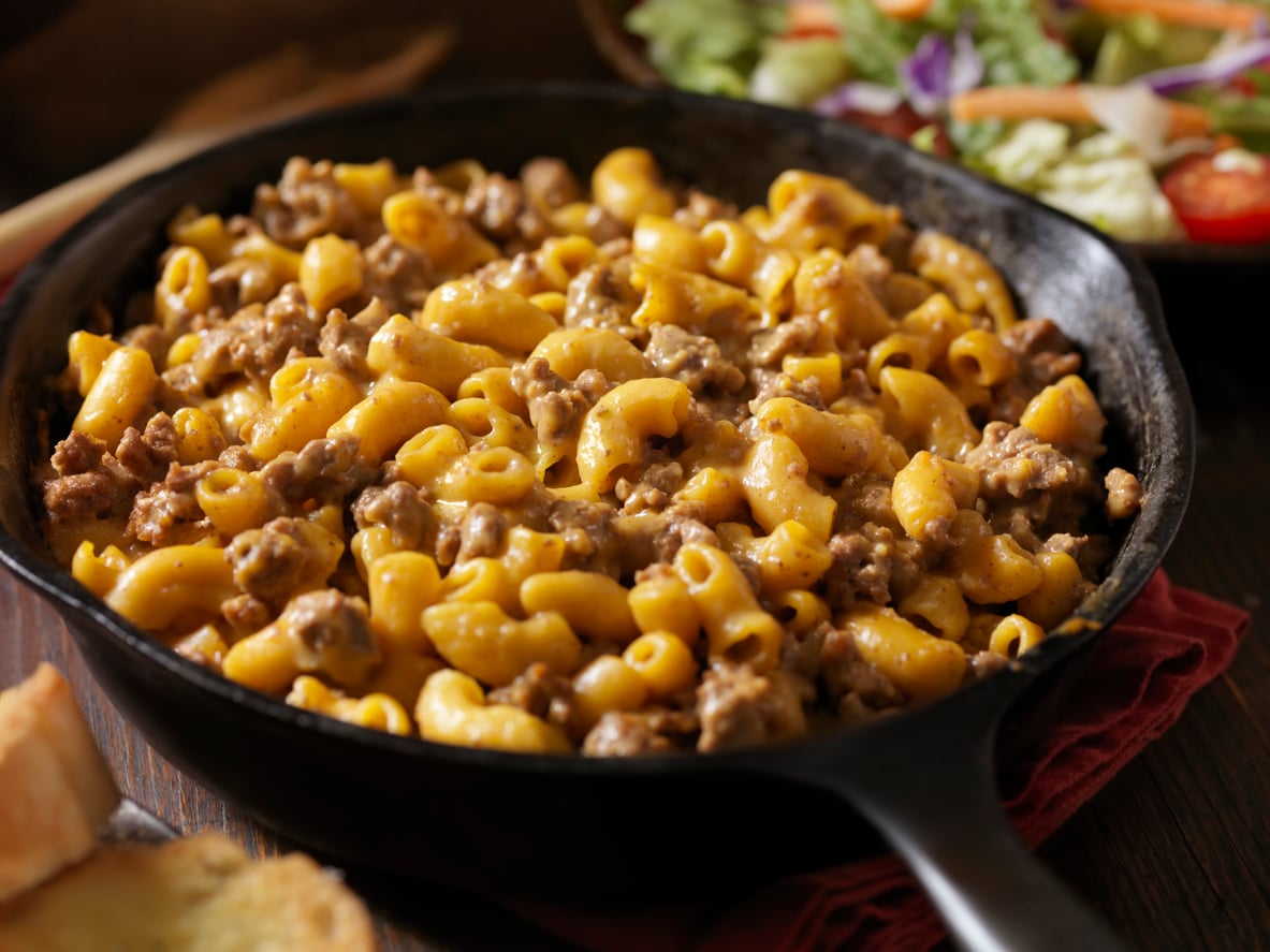 A cast iron skillet filled with macaroni and cheese mixed with ground beef, with a salad and a slice of bread visible in the background.