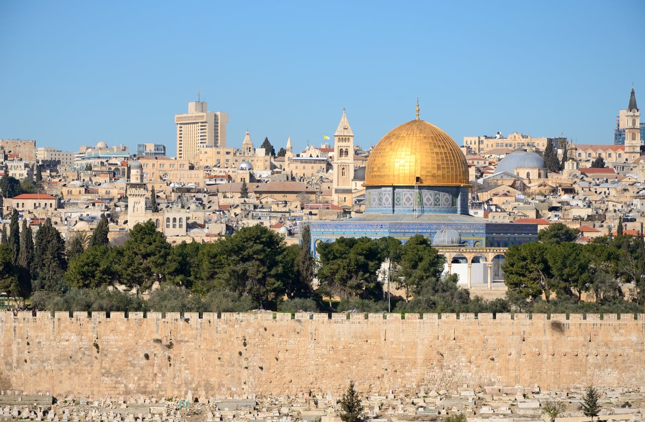 The Dome of the Rock with its golden dome is visible among historic buildings and a city wall in Jerusalem under a clear blue sky, offering a breathtaking view for visitors; however, travelers are encouraged to stay updated on current travel restrictions and advisories before planning their visit.