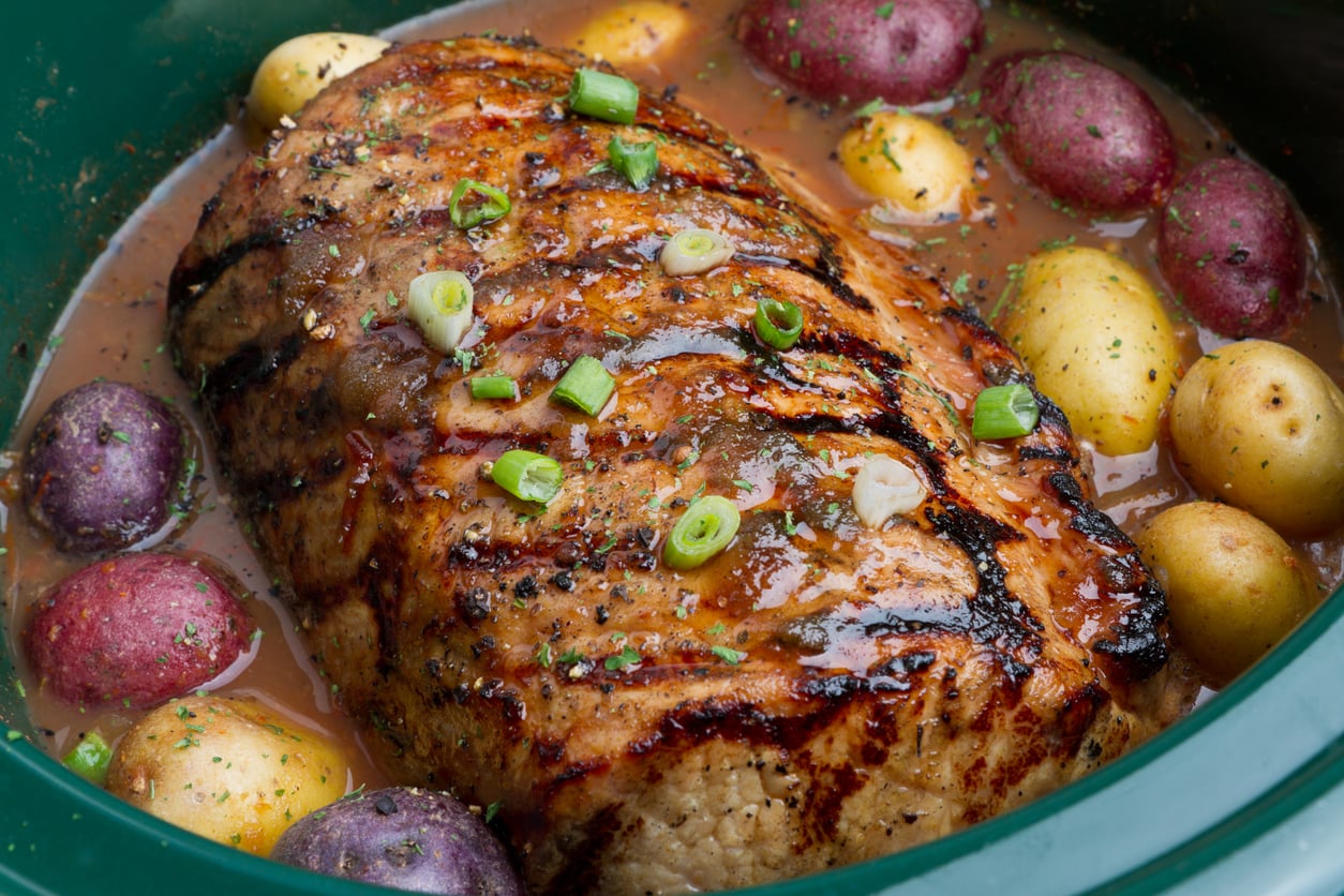 A cooked pot roast topped with sliced green onions, surrounded by red, yellow, and purple baby potatoes in a slow cooker with savory broth—just like the hearty meals middle-class moms made for Sunday family dinners.
