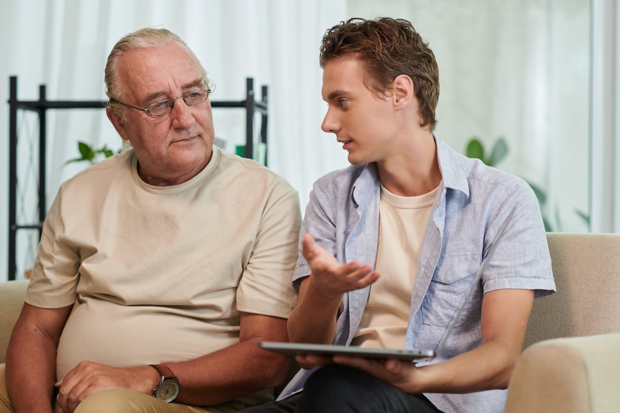 A young man holding a tablet talks to an older man sitting beside him on a couch. The older man listens attentively. Both are dressed casually in light-colored clothes.
