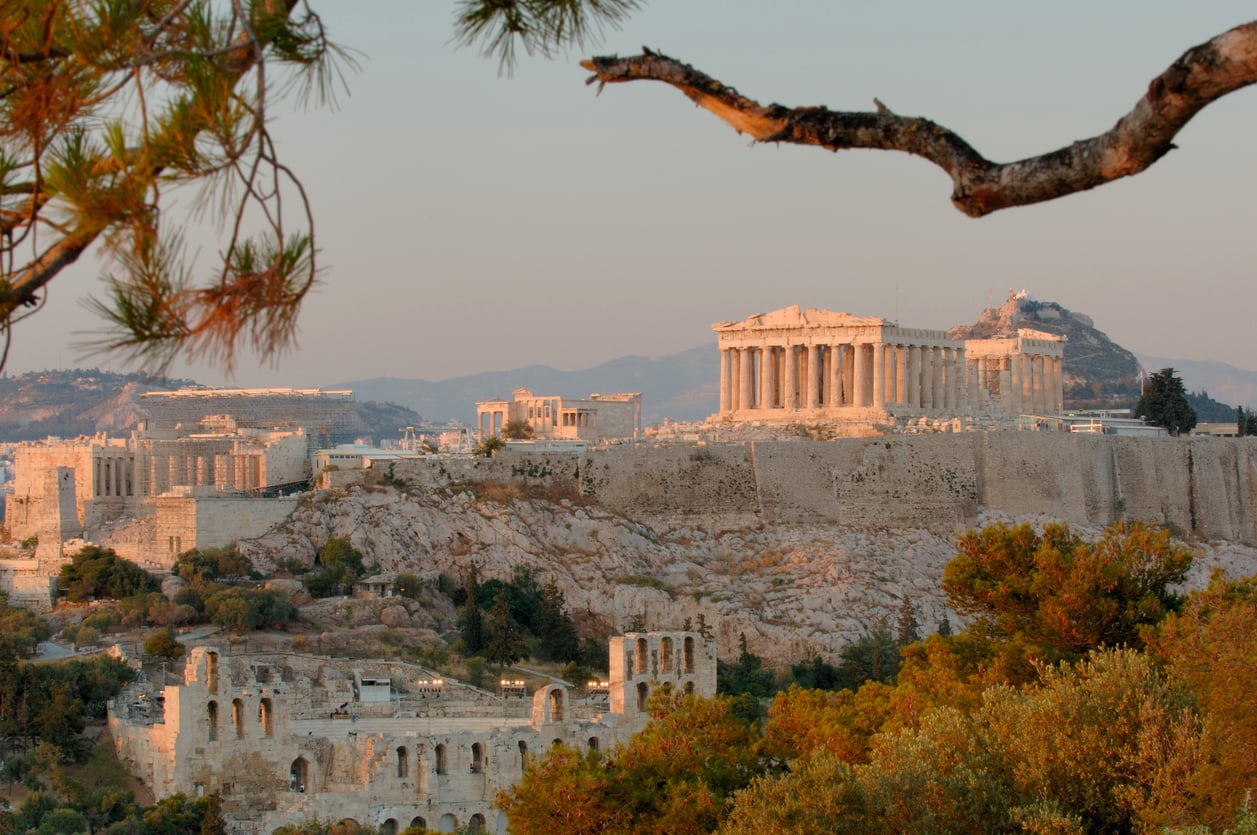 The Parthenon and other ancient ruins sit atop the Acropolis in Athens, Greece, surrounded by trees and bathed in warm evening light, with mountains in the distant background.
