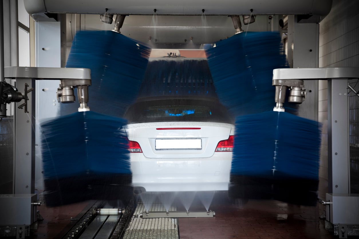 A white car is going through an automated car wash, with large blue rotating brushes cleaning its exterior and water spraying from below.