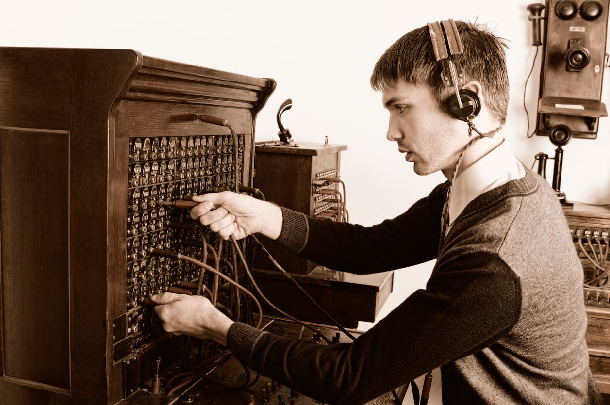 A man wearing a headset operates an old-fashioned telephone switchboard, connecting wires to different jacks—a vital piece of technology among the many things hotels used to offer guests. The scene is sepia-toned, evoking an early 20th-century office setting.