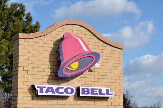 A Taco Bell sign with a pink and yellow bell logo on a brick wall, set against a blue sky with clouds and a green tree on the left&mdash;perfect for fans seeking Wild Fast-Food Facts about this iconic brand.