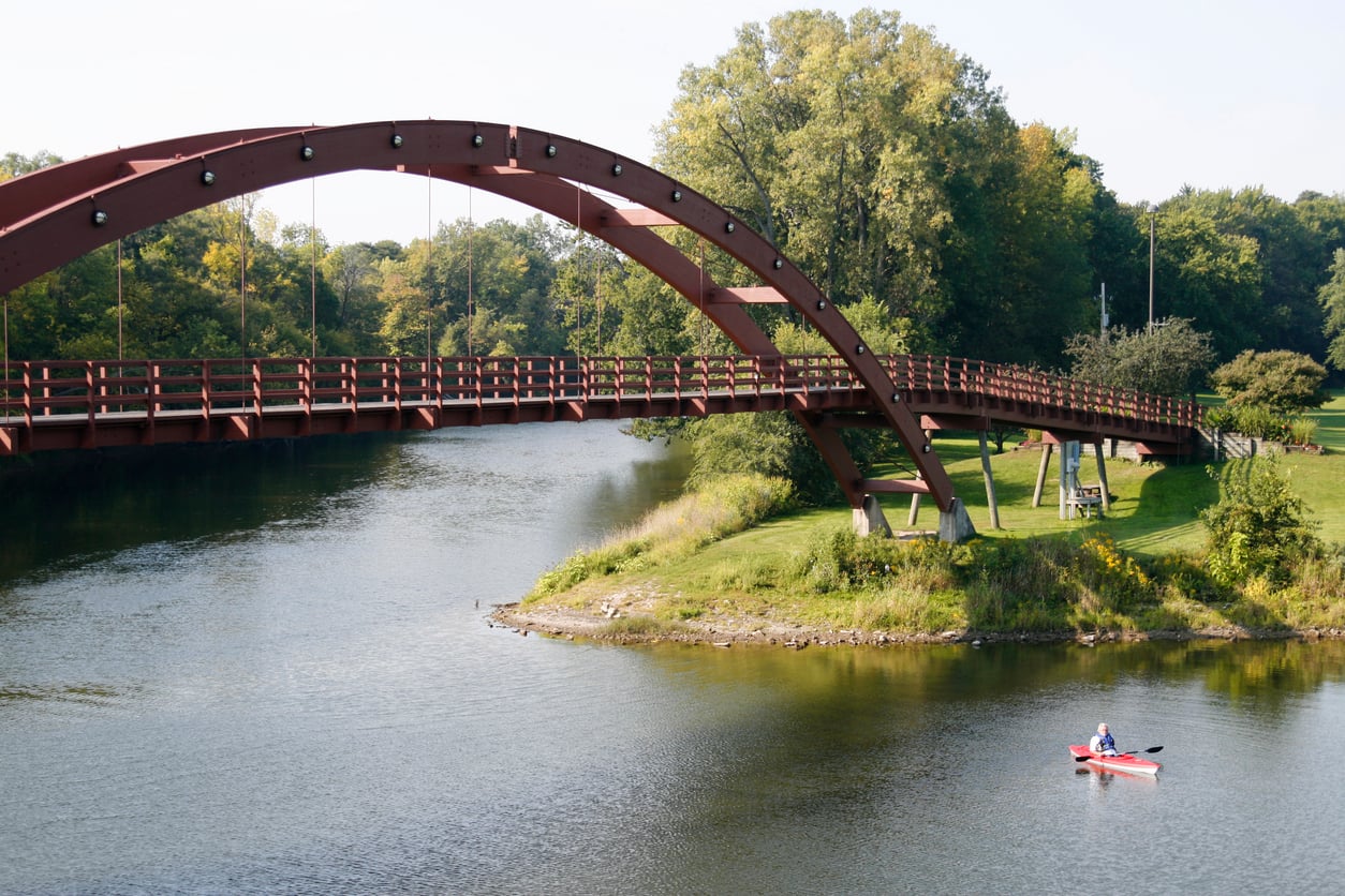 A person in a red kayak paddles on a calm river beneath a large arched pedestrian bridge, surrounded by green trees and grass. Best towns to retire in 2026