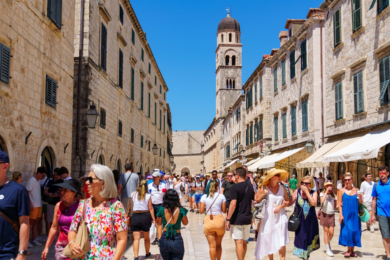 A bustling street in Dubrovnik, Croatia, with many tourists walking between historic stone buildings on a sunny day. A tall bell tower rises in the background against a clear blue sky.