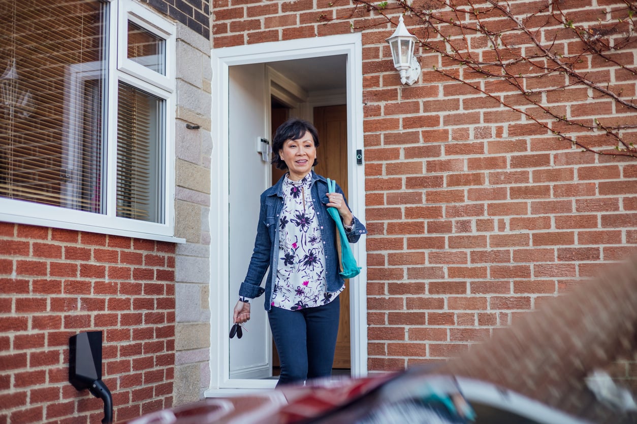 A woman with short dark hair, wearing a floral shirt and jeans, exits a house holding keys and a turquoise bag—perhaps embodying the Baby Boomer changes in lifestyle—while a brick wall and open door stand behind her.