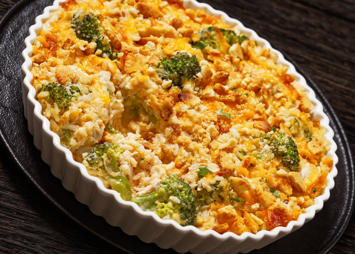 Baked broccoli casserole in a white dish, topped with a golden, crispy breadcrumb and cheese layer, sitting on a dark textured surface—a comforting classic reminiscent of the wholesome meals middle-class moms made.
