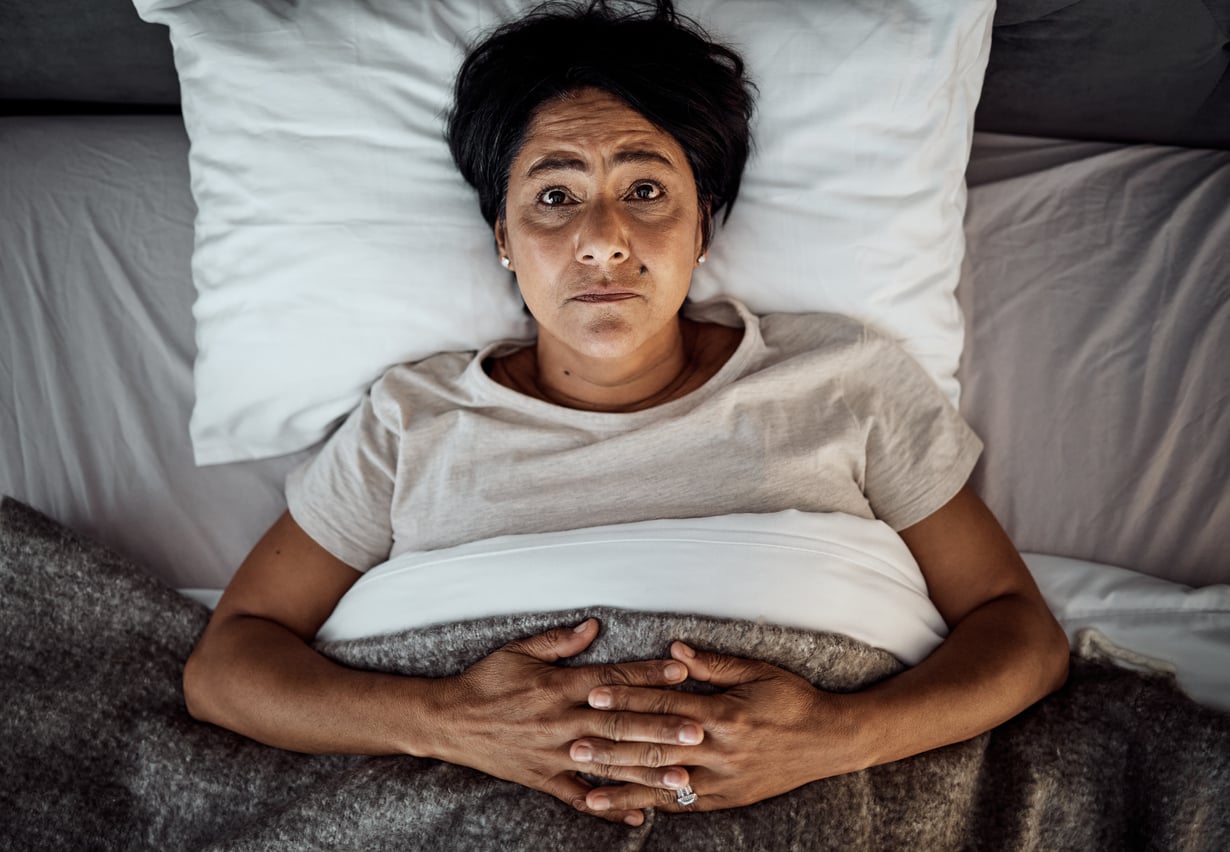 A person lies in bed on their back, looking up with a worried expression—perhaps reflecting on retirement tips or future plans. Their hands are folded on their stomach, and they are partially covered with a gray blanket. The background is a white pillow and bedding.