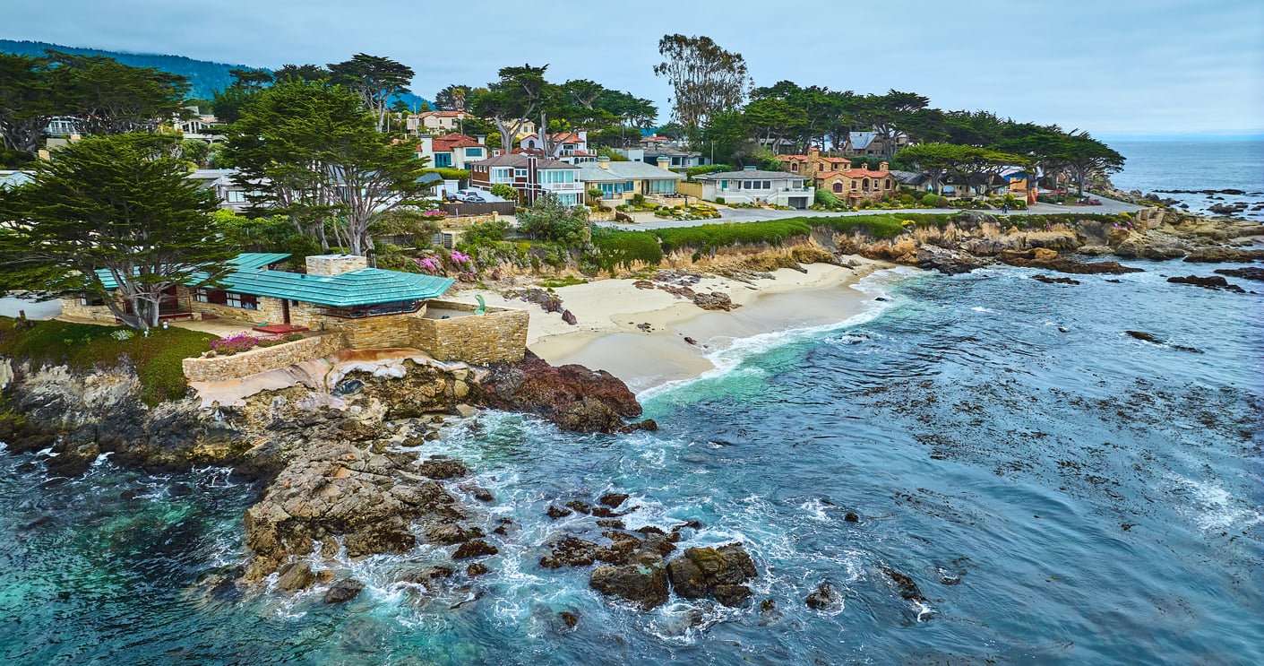 A rocky coastline with waves, sandy beaches, and houses nestled among tall trees, overlooking the ocean under a cloudy sky—an idyllic retreat perfect for relaxation and reflection after age 50.