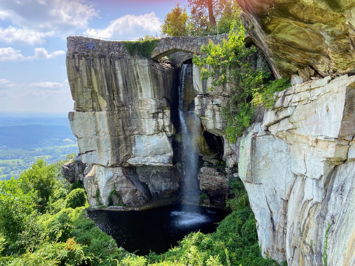 A waterfall streams over a rocky cliff into a round pool below, surrounded by lush greenery and trees, with a scenic valley and blue sky in the background—capturing the timeless charm of classic roadside attractions.