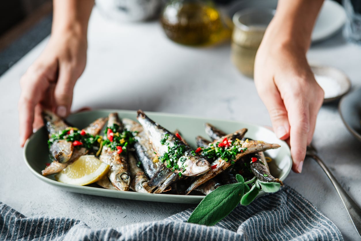 A person serves a plate of grilled sardines garnished with herbs, chopped chili, and a lemon wedge, on a striped cloth.
