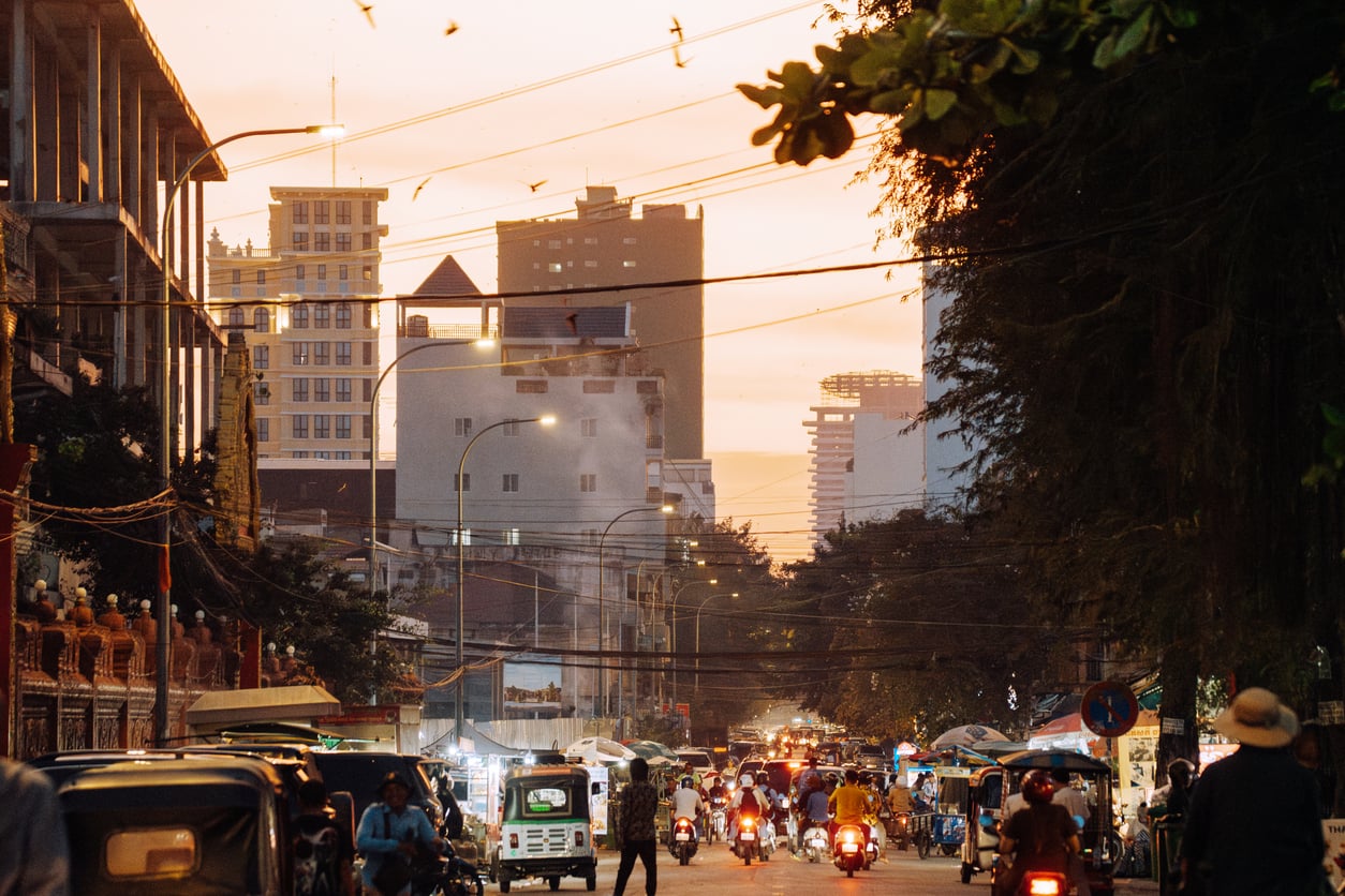 A busy city street at sunset with traffic, pedestrians, street vendors, and tall buildings in the background; despite current travel restrictions and advisories, the warm orange sky and birds flying above evoke a sense of both caution and hope.
