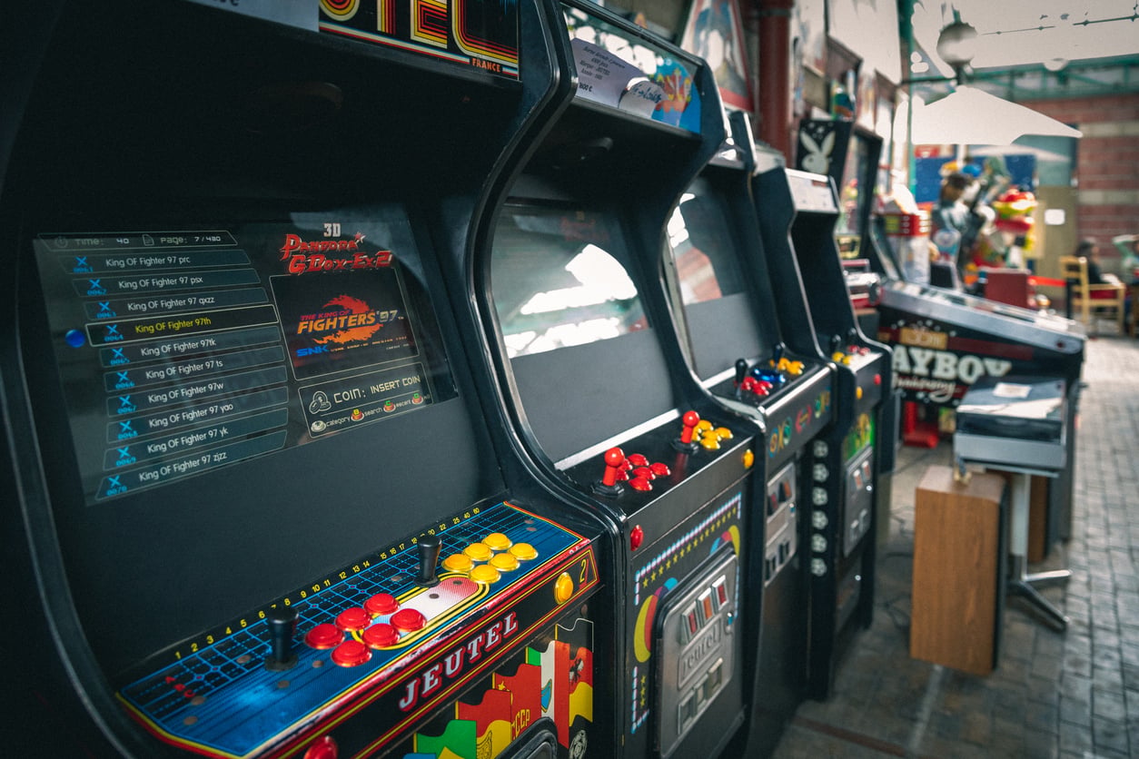 A row of vintage arcade game machines in an indoor space, with colorful buttons and joysticks. Other arcade games and seating are visible in the background.
