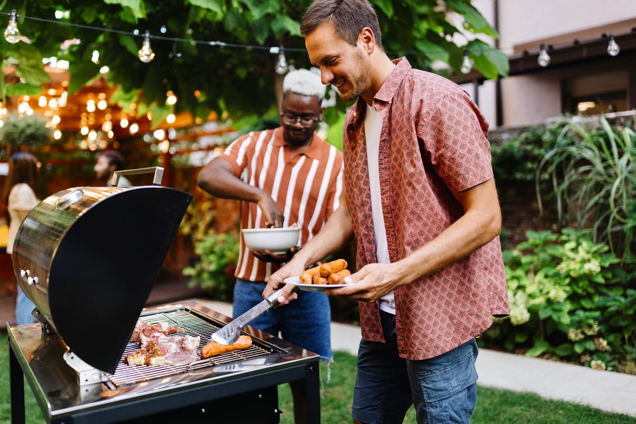 Two men grilling food outdoors at a backyard barbecue. One man is placing food on the grill, while the other prepares a bowl. String lights and greenery decorate the background.