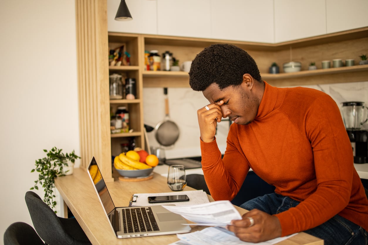 A man in an orange sweater sits at a kitchen counter, looking stressed as he reviews papers in front of his laptop, with bills, a phone, and fruit in the background.